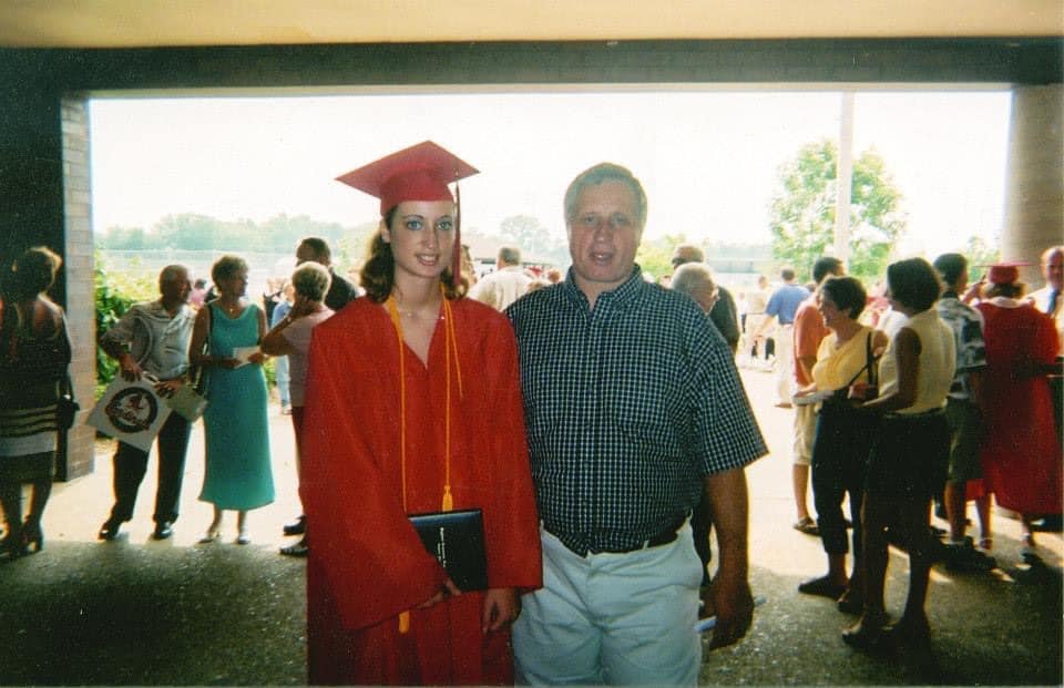 Mindy at graduation in a red graduation cap and gown standing with a smiling man.