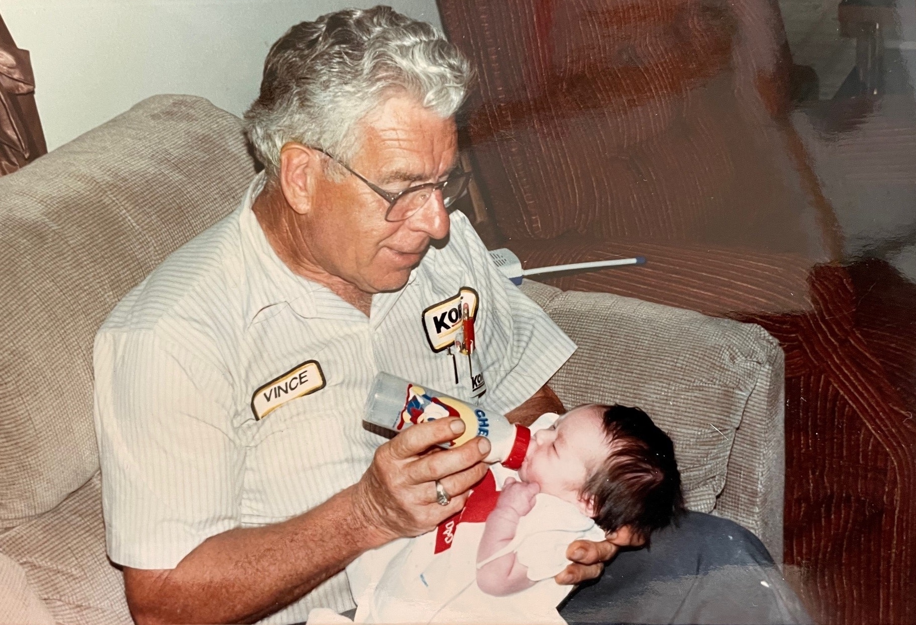 Older man happily bottle feeding a dark-haired baby.