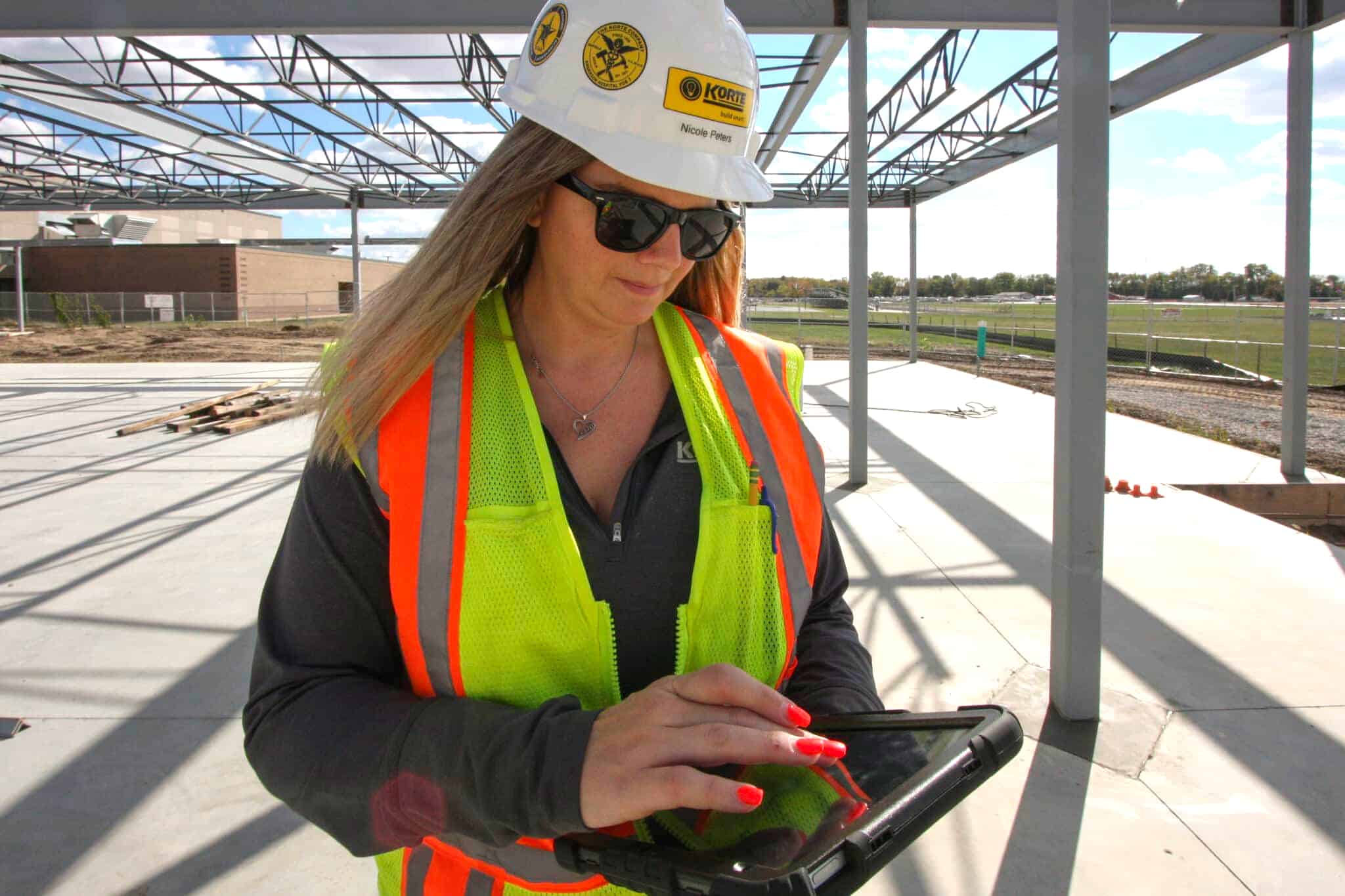 Nicole Peters in a hard hat and safety vest on an active job site with an iPad in hand. This image is meant to illustrate her role as an IT expert within a construction company.