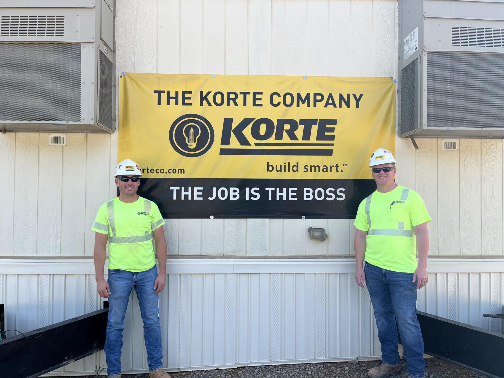 Two men in yellow safety shirts and hard hats standing on either side of a yellow Korte banner.