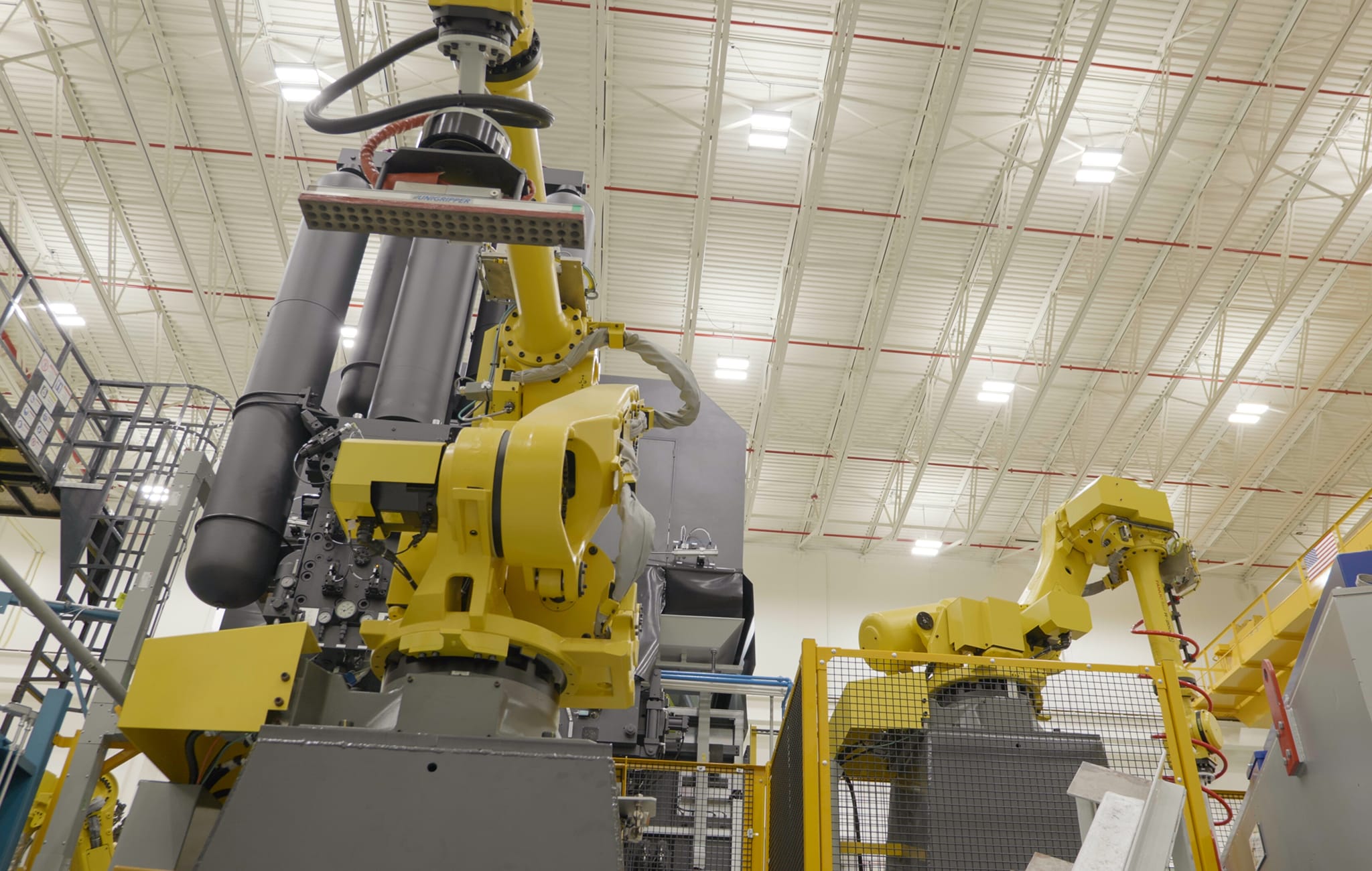 Two large yellow industrial robots with articulated arms behind safety barriers in a bright, spacious warehouse.