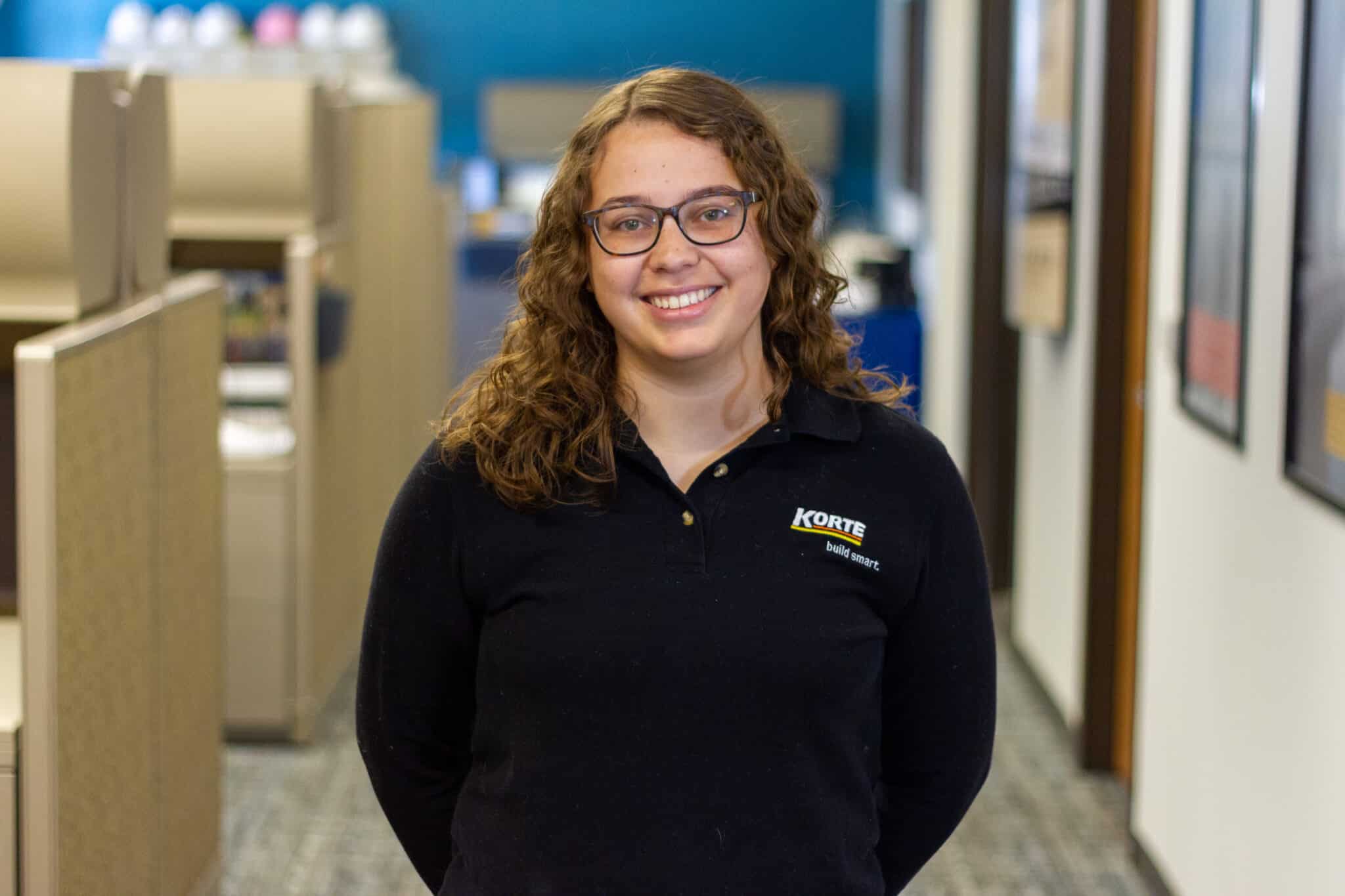 Portrait of Maggie Taylor, in the Korte office. She has glasses and a black Korte shirt on.